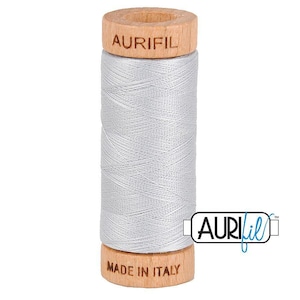 May include: A spool of light grey sewing thread wound around a wooden core. The top of the wooden spool is branded with the text AURIFIL, and the bottom is branded with the text MADE IN ITALY. A logo with the text AURIFIL is visible on the right.