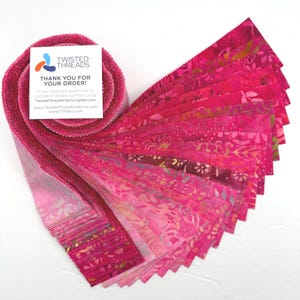 May include: A jelly roll of fabric strips in various shades of pink, magenta, and fuchsia, featuring batik-style floral and abstract patterns. A white square card with the text Twisted Threads Thank You For Your Order is placed on top.