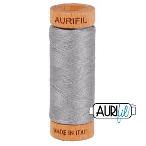 May include: A spool of gray Aurifil thread. The thread is wound around a cylindrical core with wooden ends. The top wooden end has the word "AURIFIL" printed on it. The bottom wooden end has "MADE IN ITALY" printed on it.
