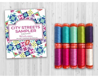 May include: A City Streets Sampler quilt pattern booklet by Jemima Flendt and a set of ten small spools of 50wt 100% cotton thread in various colors including blue, teal, green, purple, pink, magenta, and white with orange spool caps.