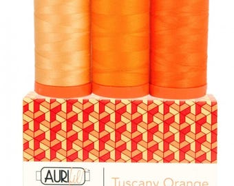 May include: Three spools of sewing thread in gradient shades of orange, ranging from light peach to vibrant orange, are positioned atop a rectangular box. The box features a geometric pattern and a label reading Aurifil Color Builder Tuscany Orange.
