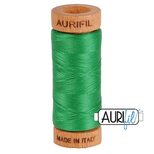 May include: A spool of vibrant green sewing thread wrapped around a light-colored wooden core. The top of the wooden spool is branded with the text AURIFIL, while the bottom base features the text MADE IN ITALY in a clear, dark font.