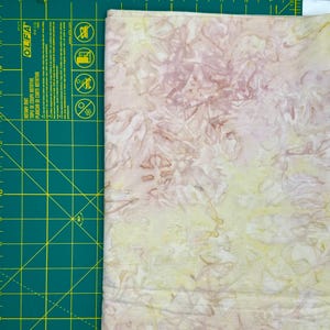 May include: Close-up of a fabric with a batik-style pattern in shades of pale yellow, pink, and brown. The fabric is folded, showing the edge. A green cutting mat with measurements in inches is visible on the left.
