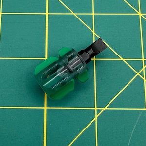 May include: A small, green and clear handled screwdriver with a black flathead tip. The handle has a clear, transparent section with green accents. The tool is designed for precision work and is set against a green cutting mat with yellow grid lines.