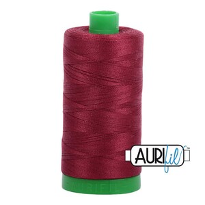 May include: A spool of deep burgundy sewing thread wound onto a cylindrical plastic core. The spool features a bright green top and bottom cap, with the brand name Aurifil printed in black and blue text on a white label at the bottom right.