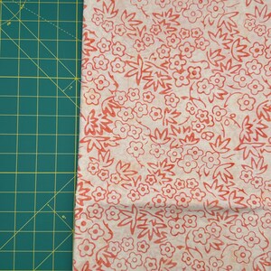 May include: Close-up of a fabric with a floral pattern. The fabric features a cream-colored background with a repeating design of red flowers and leaves. The fabric is next to a green cutting mat.