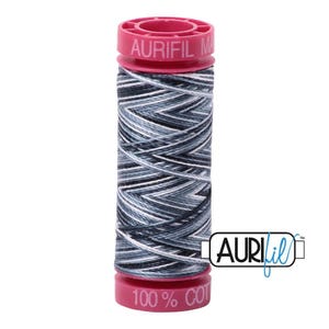 May include: A spool of Aurifil thread featuring a variegated pattern of black, charcoal, grey, and white tones. The spool has bright pink plastic ends with the text AURIFIL and 100% COT printed on them. The Aurifil logo is visible to the right.
