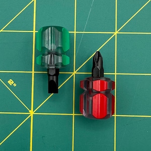 May include: Two small screwdrivers with clear plastic handles. One has a green handle and a flathead bit, the other has a red handle and a Phillips head bit. The tools are on a green cutting mat with yellow grid lines.