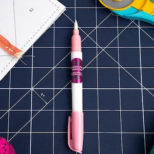 May include: A white and pink water pen with a fine tip, labeled "Foundation Paper Water Pen." The pen has a pink cap and a clip. The image also shows a 7-inch ruler, a rotary cutter, and a sewing pattern.