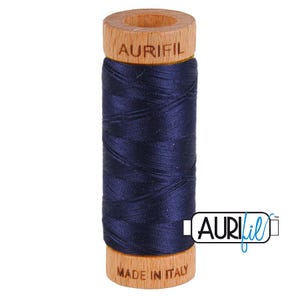 May include: A spool of dark navy blue sewing thread wrapped around a cylindrical wooden core. The top of the wooden spool is branded with the text AURIFIL, and the bottom base features the text MADE IN ITALY. The thread has a smooth texture.
