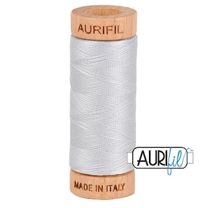 May include: A spool of light gray Aurifil thread. The thread is wound around a wooden spool with the brand name "AURIFIL" and "MADE IN ITALY" printed on the spool. The thread is tightly wound and appears smooth.