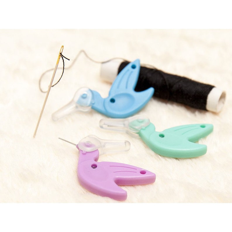 May include: Three colorful plastic threader tools shaped like birds. The threaders are blue, green, and purple. A needle and spool of black thread are also in the image.