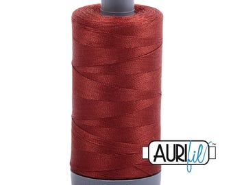 Op de afbeelding: Een cilindrische klos met diep roestkleurig naaigaren met een grijze plastic boven- en onderkant. De merknaam Aurifil is in de basis gegraveerd en een wit rechthoekig logo met de tekst Aurifil is rechtsonder op de afbeelding geplaatst.