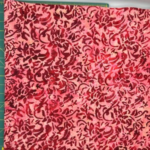 May include: A piece of fabric with a floral pattern in shades of red and pink. The design features a complex arrangement of stylized flowers and swirling shapes, creating a textured appearance. The fabric is likely cotton or a similar material.