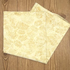 May include: A square fabric napkin featuring a light cream background with a repeating pattern of tan fern leaves. The textile has a soft, folded appearance with visible edges and a consistent botanical print across the entire surface area.