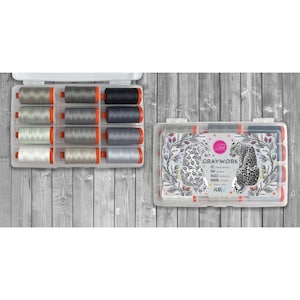 May include: A clear plastic storage case containing twelve spools of gray and black sewing thread. The lid features a decorative label with two illustrated leopards, floral accents, and the text Graywork, 12 large spools, 50 weight, 1422 yards ea, 100% cotton, Aurifil.