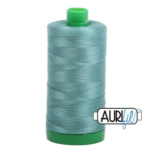 May include: A spool of teal sewing thread featuring a bright green plastic top and base. The thread is wound tightly around the cylindrical core. The brand name AURIfil is printed in black and blue text on a white rectangular label.