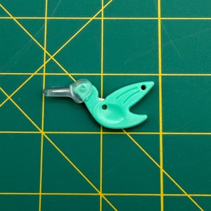 May include: A mint green bird-shaped thread cutter with a clear plastic beak cover. The tool features a small circular hole near the tail and a central opening, positioned on a green cutting mat with yellow grid lines for scale reference.