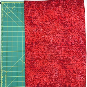May include: A piece of red fabric with a repeating leaf pattern. The fabric is next to a green cutting mat with grid lines and inch measurements. The fabric is a solid red color with darker red leaf-like shapes.