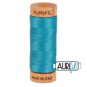 May include: A spool of vibrant turquoise sewing thread wound around a light-colored wooden core. The top of the wooden spool is branded with the text AURIFIL, and the bottom is stamped with the text MADE IN ITALY in a clear, dark font.
