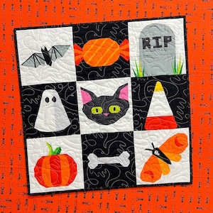 May include: A Halloween-themed quilt with nine squares, each featuring a different applique design. The designs include a bat, candy, tombstone, ghost, black cat, candy corn, pumpkin, bone, and a moth. The quilt is bordered by an orange fabric with skeleton figures.