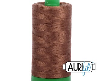 Puede incluir: Un carrete cilíndrico de hilo de coser marrón con una parte superior y una base de plástico verde brillante. El carrete está envuelto con hilo marrón oscuro liso y muestra el nombre de la marca AURIfil en un logo blanco rectangular.