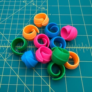 May include: A collection of colorful plastic thread holders in various shades of blue, green, pink, and orange. The holders are shaped like a spiral and are designed to hold thread for sewing or crafting.