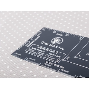 May include: A black cross-stitch key with white text and measurements. The key includes a ruler, reference guide for floss strands and needle sizes, and a cloth counter. The background is a light gray fabric with white polka dots.