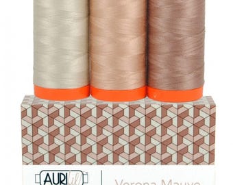 Op de afbeelding: Drie klossen garen in verschillende tinten beige, bruin en mauve, elk voorzien van feloranje plastic doppen aan de boven- en onderkant. De klossen rusten op een rechthoekige doos met een geometrisch patroon en het label Aurifil Color Builder.