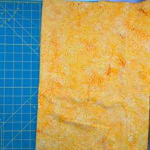 May include: A piece of fabric with a vibrant yellow and orange floral pattern. The design features stylized flower-like shapes in varying shades of yellow and orange. The fabric is next to a blue cutting mat with grid lines.
