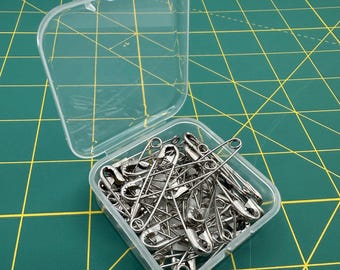 May include: A clear plastic container filled with silver safety pins. The container is open, revealing the pins inside. The background is a green cutting mat with yellow grid lines. The safety pins are made of metal.