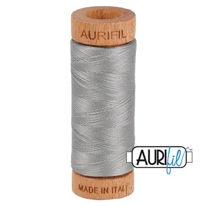 May include: A spool of silver-grey sewing thread wound around a cylindrical wooden core. The top of the wooden spool is embossed with the brand name AURIFIL, and the bottom is embossed with the text MADE IN ITALY. The thread is smooth.