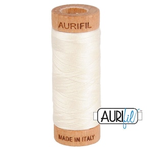 May include: A spool of off-white sewing thread wound around a cylindrical wooden core. The top of the wooden spool is branded with the text AURIFIL, and the bottom is branded with the text MADE IN ITALY. The thread has a smooth, matte finish.