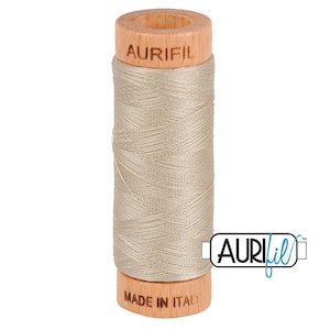 May include: A spool of light beige sewing thread wound around a cylindrical wooden core. The top of the wooden spool is branded with the text AURIFIL, and the bottom is branded with the text MADE IN ITALY. The thread has a smooth, matte finish.