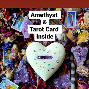 May include: A white heart-shaped box with a purple amethyst crystal inside. The box is surrounded by dried flowers and a tarot card with the text "The Hermit".