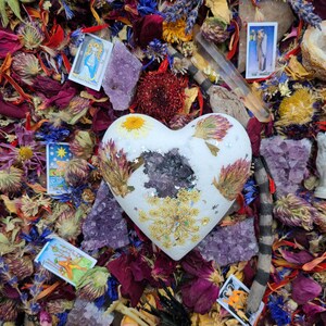 May include: A white heart-shaped object decorated with dried flowers, gold accents, and a dark center, surrounded by amethyst crystals, tarot cards, and colorful dried flowers. A clear crystal wand is also present.