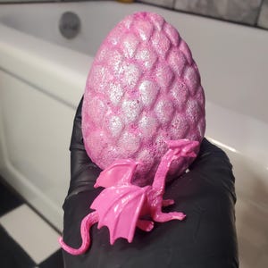 Puede incluir: Una bomba de baño rosa en forma de huevo con una superficie texturizada y una pequeña figura de dragón rosa. La bomba de baño tiene un brillo brillante. El dragón está posado junto a la bomba de baño. El fondo incluye una bañera.