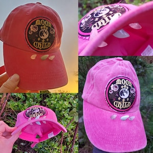 May include: Pink baseball cap with a round embroidered patch that says "Moon Child" with a gold and black design. The cap has white crystals attached to the brim.