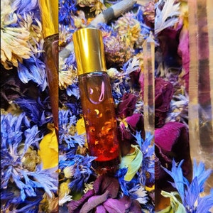May include: A small amber glass roller bottle with a gold lid and a gold cap. The bottle is filled with a liquid that has a glittery, golden appearance. The bottle is surrounded by dried flowers and leaves.