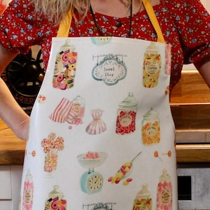 May include: A white apron with a yellow trim and a colorful pattern of candy jars, lollipops, and a sign that says "Sweet Shop".