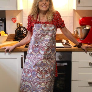May include: A woman wearing a red floral print apron with a white background. The apron has a long, flowing design and features a variety of floral patterns. The woman is standing in a kitchen with a stand mixer and other kitchen appliances in the background.