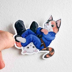 Puede incluir: Una pegatina con dos gatos en un combate de jiu-jitsu. El gato de arriba lleva un gi azul y tiene un patrón calico. El gato de abajo es blanco y tiene la nariz negra.