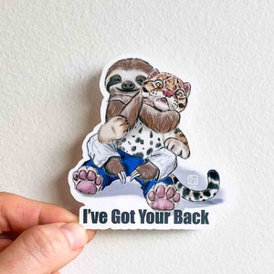 Könnte beinhalten: Ein Aufkleber mit einer Cartoon-Illustration eines Faultiers und eines Ozelots, die einen blauen und weißen Gi tragen. Der Ozelot hat seinen Arm um den Hals des Faultiers gelegt und hält den Arm des Faultiers fest. Der Text "I've Got Your Back" ist unter dem Bild gedruckt.