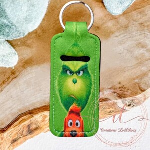 May include: Green Grinch-themed keyring with a silver ring. The keyring features a cartoon image of the Grinch and a red character. The text "Créations LouChoux" is printed on the bottom right corner.