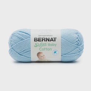 Könnte beinhalten: Ein Knäuel hellblaues Garn mit einem weißen Papieretikett, das den Text Yarnspirations BERNAT Softee Baby Cotton, eine Baby-Illustration und ein kreisförmiges Siegel zeigt. Das Etikett gibt ein Gewicht von 120 g oder 4,2 oz an.
