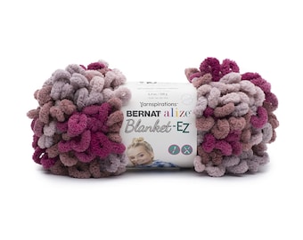 Puede incluir: Una madeja de hilo Bernat Alize Blanket-EZ con una paleta de colores variados en magenta intenso, malva y lavanda claro. El hilo tiene una textura de bucles preformados para tejer con los dedos. La etiqueta muestra 180 g de peso.