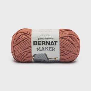 Può includere: Un gomitolo di filato Bernat Maker color terracotta con un'etichetta di carta bianca. L'etichetta riporta il testo Yarnspirations, Bernat e Maker, insieme al peso di 250 g e 8,8 oz, e un piccolo grafico di un capo lavorato su una sedia.