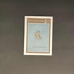 Puede incluir: Un botón de media luna pintado a mano con un fondo marrón claro y detalles morados. El botón está en una tarjeta con el texto "Lynette Anderson Designs" y "Hand Painted in Australia".
