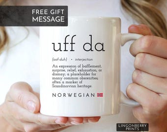 May include: White ceramic mug with the text "uff da" in black, along with a definition and the word "NORWEGIAN" and a Norwegian flag. The mug is held by a person.