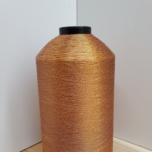 May include: A large spool of metallic copper-colored thread. The thread is tightly wound around a cylindrical core with a black base and top. The thread has a textured appearance, reflecting light.